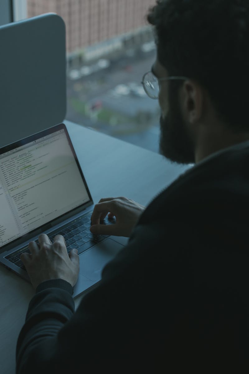 A man coding on his laptop by a window in an office setting, showcasing technology work.